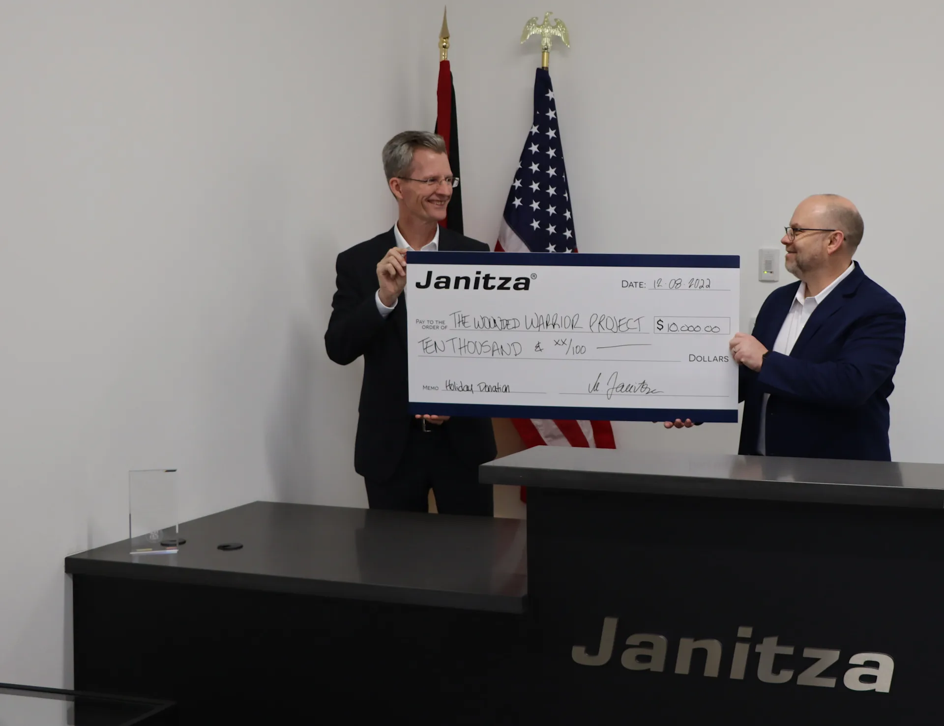 Übergabe eines großen Schecks durch Janitza-Mitarbeiter in einem Büro mit amerikanischen Flaggen im Hintergrund.