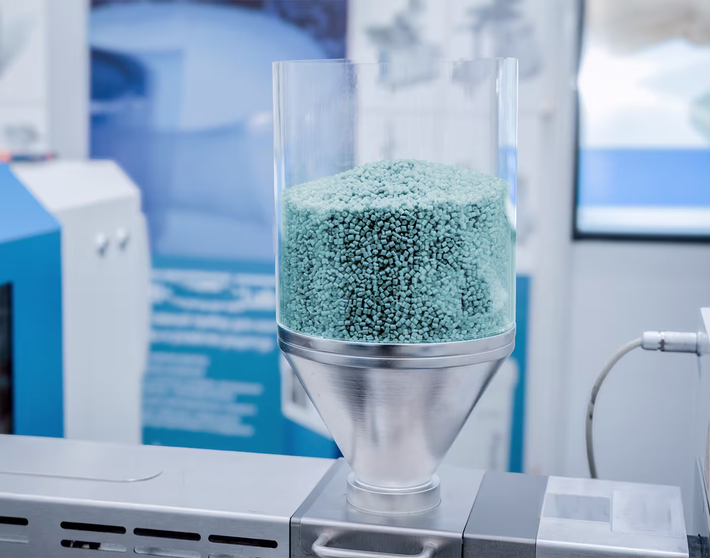 Ein transparenter Behälter mit hellblauen Kunststoffgranulaten steht auf einer metallischen Maschine in einer industriellen Umgebung.