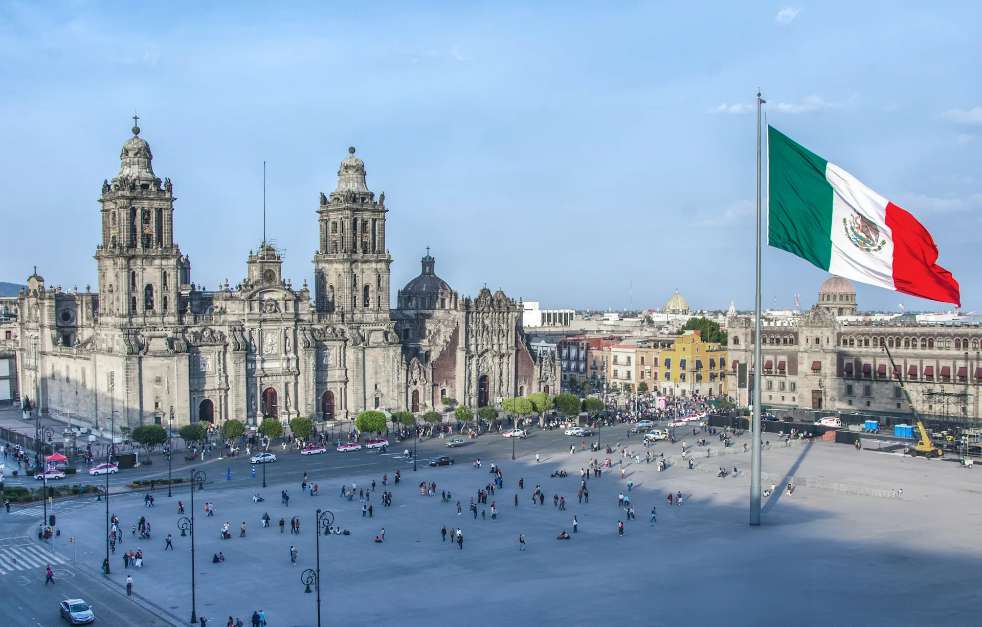 Mexico City's Zócalo square with Metropolitan Cathedral, bustling crowd, and large Mexican flag waving in foreground.
